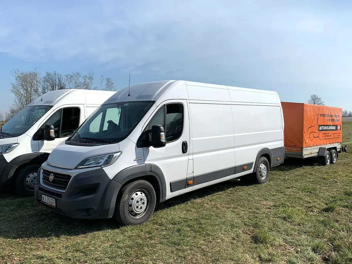 Biały Fiat Ducato z pomarańczową przyczepą na trawiastym placu
