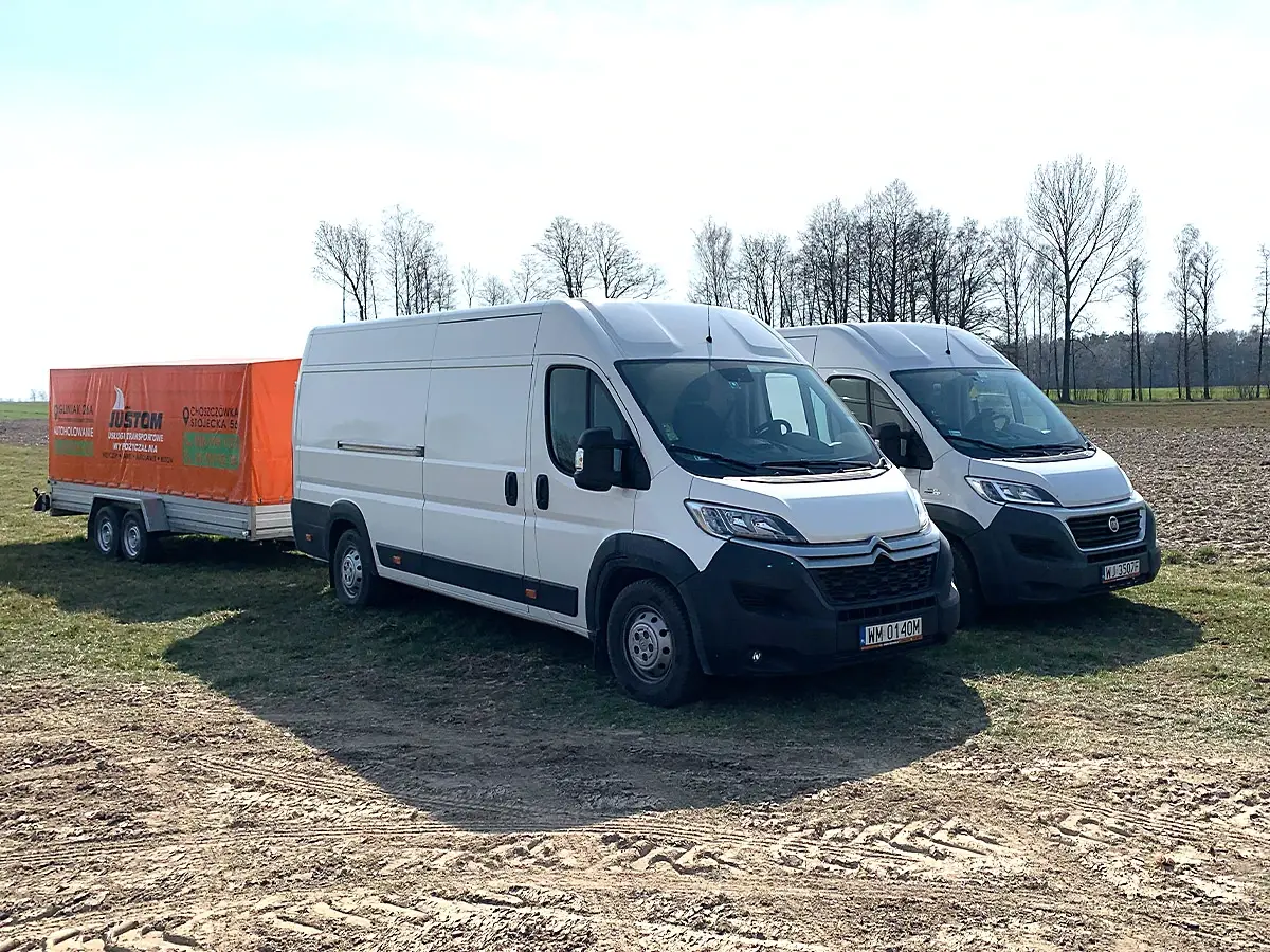Dwa białe busy dostawcze z pomarańczową przyczepą na polu
